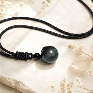 Pendentif Obsidienne œil céleste avec cordon noir posé sur fond minéral clair et décor végétal